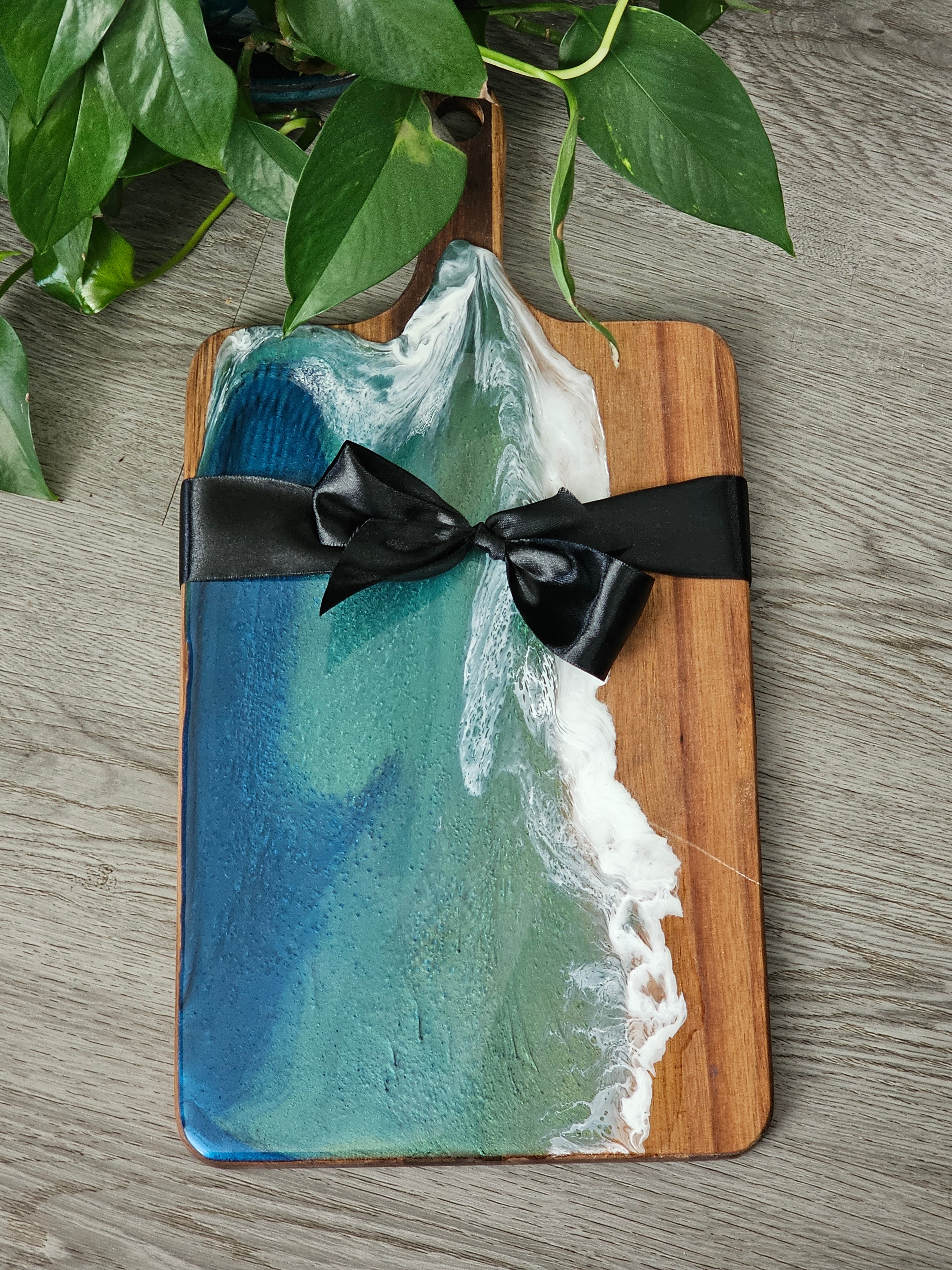 Wooden cutting board with ocean wave design and black ribbon on a wooden surface.